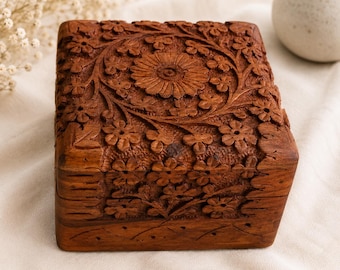 Puede incluir: Una caja cuadrada de madera con intrincados patrones florales tallados a mano en la tapa y el panel frontal. La madera de color marrón oscuro muestra una veta natural, con una gran flor central rodeada de pequeñas flores y enredaderas.