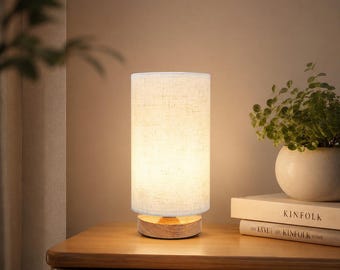 May include: A cylindrical table lamp with a textured off-white fabric shade and a round light-toned wooden base. The lamp emits a warm, soft glow, illuminating the surrounding area with a gentle light, creating a calm and inviting atmosphere.