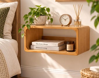 May include: A light wood floating wall shelf with rounded corners. On top sits a potted green plant, a round analog clock, and a reed diffuser. Inside the shelf are two stacked books titled The Cozy Home and Minimal Living, and a brown candle.