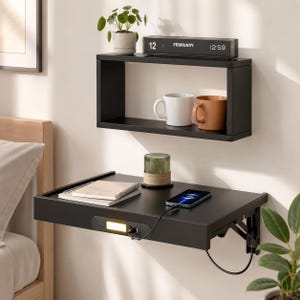 May include: A black wall-mounted floating shelf with a rectangular frame and a separate black folding desk shelf below. The desk features a built-in light, a power outlet, and a charging cable. The shelf holds a digital clock, mugs, and a plant.