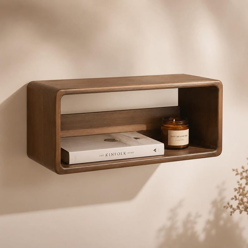 Puede incluir: Un estante de pared rectangular de madera con esquinas redondeadas y acabado marr&oacute;n oscuro. Dentro del estante hay un libro blanco titulado The Kinfolk Home y un peque&ntilde;o frasco de vela de vidrio con etiqueta marr&oacute;n y una llama encendida.
