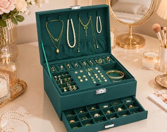 Puede incluir: Un joyero de terciopelo verde oscuro con tapa abatible y un cajón extraíble. El interior cuenta con compartimentos para collares, pendientes y anillos, con varios collares dorados colgados de ganchos y diversos anillos guardados en el cajón.