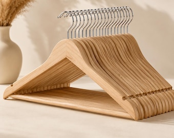 Puede incluir: Un juego de perchas de madera de color claro con un acabado de veta de madera natural suave y ganchos giratorios de metal plateado pulido. Cada percha tiene una barra horizontal para pantalones y muescas para asegurar las prendas.
