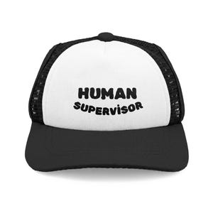 May include: A black and white trucker hat with the words "HUMAN SUPERVISOR" printed in bold, black letters across the front. The hat features a black brim and mesh back, with a white front panel.