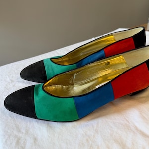 May include: A pair of low-heeled pumps featuring a color-blocked design with a black suede toe cap, followed by panels of green, blue, and red suede. The interior lining is a metallic gold finish with a low, rounded heel and a closed toe.