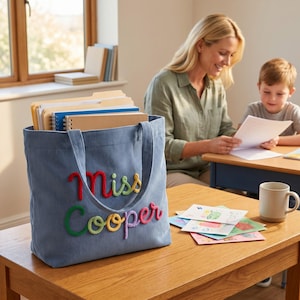 Puede incluir: Un bolso tote de pana azul claro con el texto "miss Cooper" bordado en letras de hilo en relieve de varios colores. El bolso está lleno de varios cuadernos y carpetas, y se encuentra sobre una mesa de madera junto a una taza de cerámica blanca.