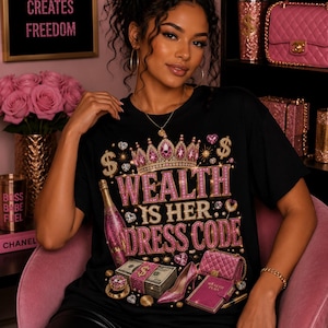 May include: A black t-shirt featuring a graphic print with the text WEALTH IS HER DRESS CODE in pink and gold letters. The design includes a jeweled crown, a bottle of pink champagne, stacks of cash, a pink high heel shoe, and a pink quilted handbag.