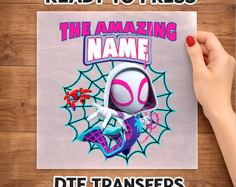 Op de afbeelding: Een DTF-transfervel met een stripfiguur in een wit, paars en blauw pak met capuchon, geplaatst voor een zwarte spinnenwebafbeelding. De tekst THE AMAZING NAME staat in roze en wit boven de figuur gedrukt.