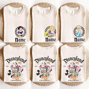 Puede incluir: Camisetas de color crema con gr&aacute;ficos de personajes de Disney, incluyendo Blancanieves, Rapunzel y Elsa, cada uno en un c&iacute;rculo a cuadros con la palabra "Name" debajo. Las camisetas inferiores presentan un gr&aacute;fico de Disneyland con el texto "Est 1955 CALIFORNIA."