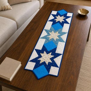 May include: A rectangular quilted table runner featuring a repeating star pattern in shades of navy blue, bright blue, and cream. The design includes three large star motifs with intricate stitching, bordered by white fabric panels.
