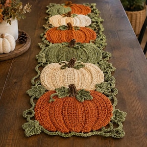May include: A crochet table runner featuring a repeating pattern of pumpkins in shades of orange, cream, and sage green, accented with dark brown stems and green crocheted leaves. The runner has a textured, ribbed design throughout the pumpkins.