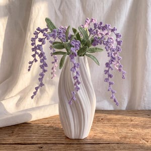 May include: A white ceramic vase with a spiraling, twisted design holds a bouquet of handcrafted crochet lavender stems. The purple crochet flowers feature intricate, textured petals and are accompanied by detailed, light green crocheted leaves.