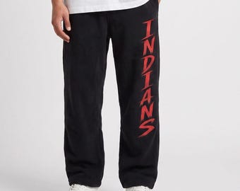 May include: A plain white short-sleeved t-shirt paired with black sweatpants. The sweatpants feature the word INDIANS printed vertically in a stylized red font along the left leg. The outfit is completed with white athletic sneakers.