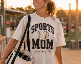 May include: A cream-colored short-sleeve t-shirt with black text that reads SPORTS MOM and Game Day Vibes, featuring a baseball graphic in the shape of a heart. The shirt is paired with a black baseball cap, black leggings, and white sneakers.