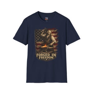 May include: Navy blue t-shirt featuring a graphic of a blacksmith with the American flag in the background. The text "FORGED IN FREEDOM" is displayed below the image. The shirt has a crew neck and short sleeves.