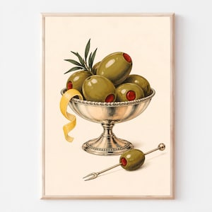 Op de afbeelding: Een ingelijste kunstdruk met een zilveren schaal op voet gevuld met groene olijven gevuld met rode piment. Een gele citroenschil hangt over de rand van de schaal en een enkele olijf op een zilveren cocktailprikker ligt onder de schaal.