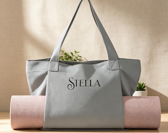 May include: A light gray canvas tote bag featuring the name Stella printed in black serif font on the front. The bag has two shoulder straps and a horizontal opening at the base, which holds a rolled-up light pink yoga mat with a grid texture.