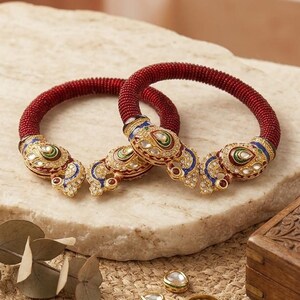 Könnte beinhalten: Ein Paar dunkelrote Perlenarmreifen mit verzierten goldfarbenen Metallenden, die mit buntem Emaille und klaren Steinen besetzt sind. Ein kleines weißes Etikett mit der Aufschrift Handcrafted Kundan Bangle Pair liegt neben losen Steinen.