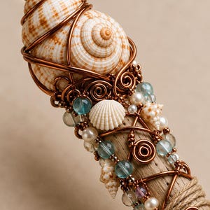 May include: A decorative wand featuring a large, striped seashell wrapped with copper wire. The wand is adorned with small seashells, blue and clear beads, and pearls. The base is wrapped with twine.