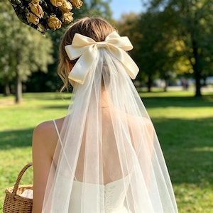Könnte beinhalten: Rückansicht einer Braut in weißem Hochzeitskleid und Schleier. Eine große, cremefarbene Satinschleife ist am Hinterkopf der Braut befestigt, mit einem langen, durchsichtigen Schleier. Der Hintergrund ist ein grüner Park.