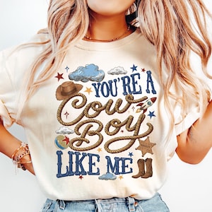 K&ouml;nnte beinhalten: Ein cremefarbenes Kurzarm-T-Shirt mit einem Grafikdruck, der den Text "YOU'RE A COWBOY LIKE ME" in Blau und Braun zeigt. Das Design enth&auml;lt Illustrationen eines braunen Cowboyhuts, eines Seils, einer Rakete, eines Sheriffsterns und Stiefeln.