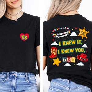 May include: A black t-shirt featuring a small red heart on the front with the letters TS. The back displays a colorful graphic with the text I KNEW IT, I KNEW YOU, TS5, and INFINITY AND BEYOND, alongside illustrations of cowboy boots, a hat, and a star.