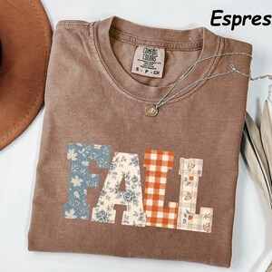 May include: An espresso-coloured t-shirt with a crew neckline and a graphic print on the front that spells the word FALL. Each letter features a different pattern, including blue floral, cream floral, orange gingham, and a plaid design with small flowers.