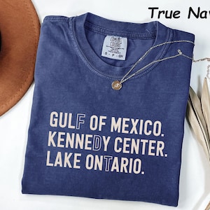 May include: A True Navy short-sleeve t-shirt with a crew neckline. The shirt features the text GULF OF MEXICO. KENNEDY CENTER. LAKE ONTARIO. printed in a light beige color. The Comfort Colors brand label is visible at the inner neck area.