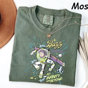 May include: Moss green t-shirt featuring a graphic of Buzz Lightyear with the text "Buzz Lightyear" and "To Infinity and Beyond." The shirt has a crew neck and short sleeves. A gold necklace is draped across the shirt.