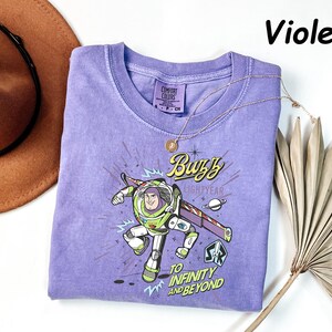 May include: A violet Comfort Colors t-shirt featuring a graphic of Buzz Lightyear with the text "Buzz Lightyear" and "To Infinity and Beyond." A gold necklace and a brown hat are also in the image.