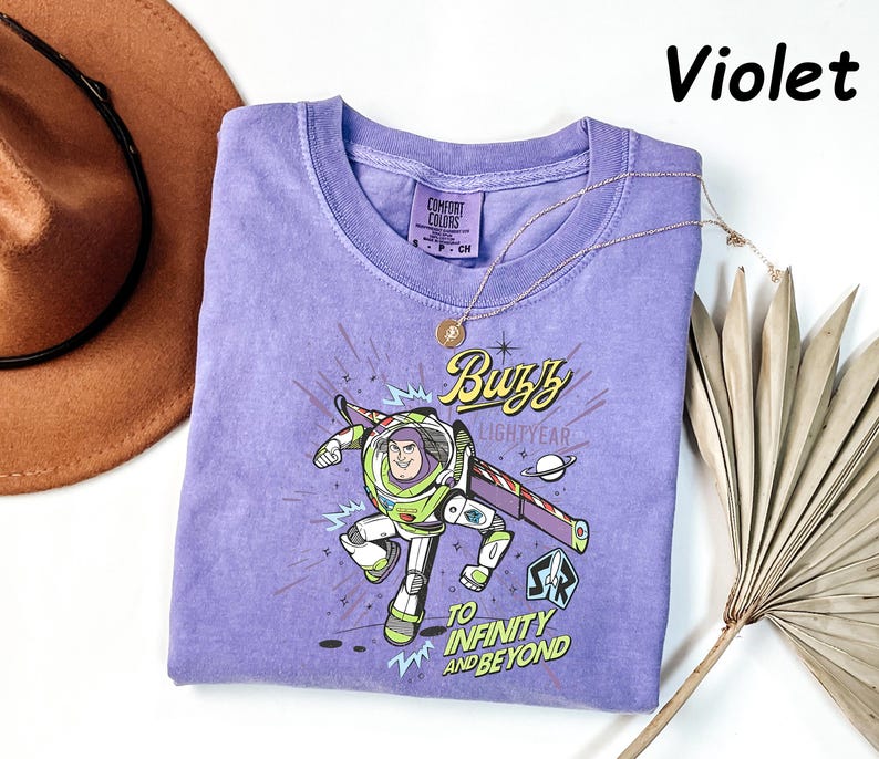 May include: A violet Comfort Colors t-shirt featuring a graphic of Buzz Lightyear with the text "Buzz Lightyear" and "To Infinity and Beyond." A gold necklace and a brown hat are also in the image.