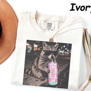 May include: Ivory-colored t-shirt featuring a raccoon wearing sunglasses and drinking from a can. The can has a pink and blue design with the word "Fluffy". A gold necklace and a brown hat are also visible.