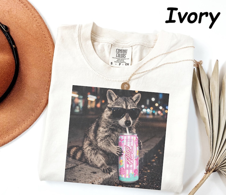 May include: Ivory-colored t-shirt featuring a raccoon wearing sunglasses and drinking from a can. The can has a pink and blue design with the word "Fluffy". A gold necklace and a brown hat are also visible.