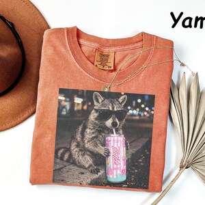 May include: A coral-colored t-shirt featuring a graphic of a raccoon wearing sunglasses and drinking from a can. The can has a pink and blue floral design. A gold necklace and a brown hat are also in the image.