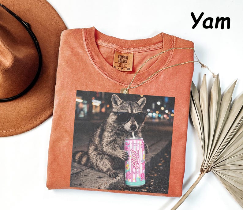 May include: A coral-colored t-shirt featuring a graphic of a raccoon wearing sunglasses and drinking from a can. The can has a pink and blue floral design. A gold necklace and a brown hat are also in the image.