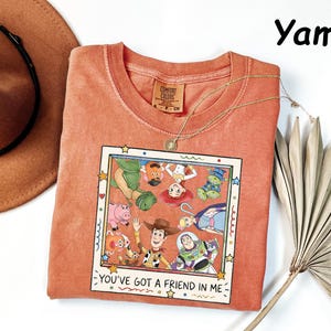 May include: A yam-colored t-shirt featuring a square graphic print of Toy Story characters including Woody, Buzz Lightyear, and Jessie. The print is framed like a Polaroid photo with the text "YOU'VE GOT A FRIEND IN ME" printed at the bottom.