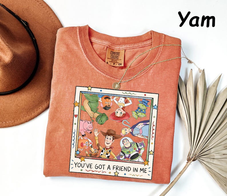 May include: A yam-colored t-shirt featuring a square graphic print of Toy Story characters including Woody, Buzz Lightyear, and Jessie. The print is framed like a Polaroid photo with the text "YOU'VE GOT A FRIEND IN ME" printed at the bottom.