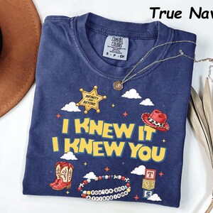 May include: A true navy blue short-sleeve t-shirt featuring a graphic print with the text I KNEW IT I KNEW YOU, a yellow sheriff star badge with the text INFINITY AND BEYOND, a red cowboy hat, red cowboy boots, and a beaded bracelet with text.