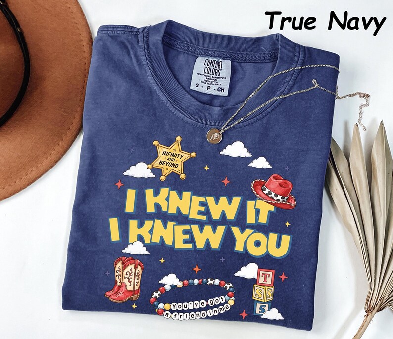 May include: A true navy blue short-sleeve t-shirt featuring a graphic print with the text I KNEW IT I KNEW YOU, a yellow sheriff star badge with the text INFINITY AND BEYOND, a red cowboy hat, red cowboy boots, and a beaded bracelet with text.