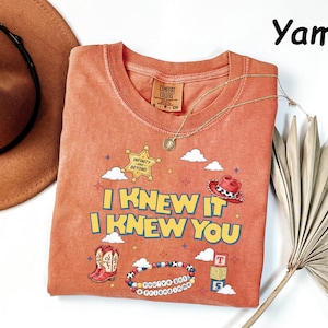 May include: A rust-colored t-shirt featuring a graphic print with the text I KNEW IT I KNEW YOU, a sheriff star badge with INFINITY AND BEYOND, a red cowboy hat, cowboy boots, a beaded bracelet with YOU'VE GOT A FRIEND IN ME, and toy blocks.