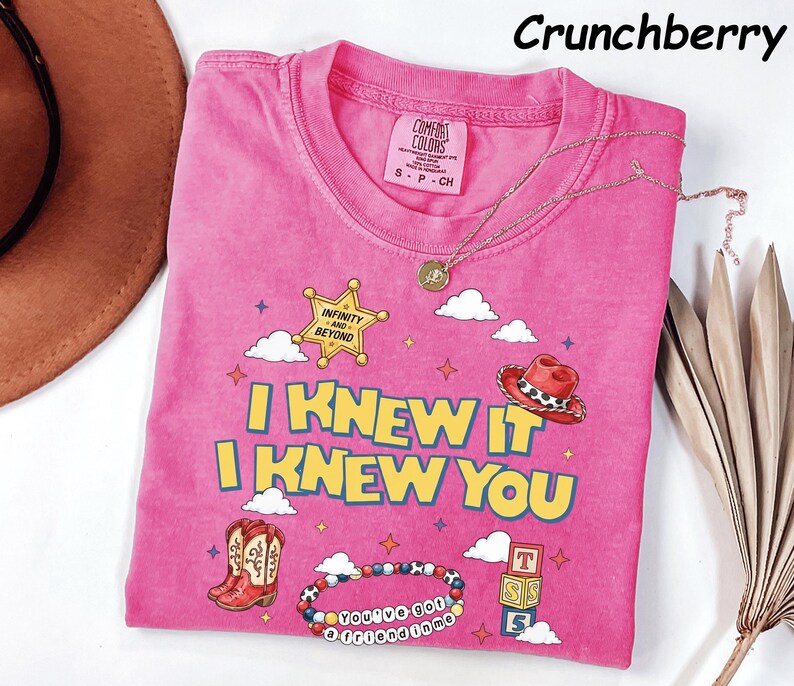 May include: A pink short-sleeve t-shirt featuring a graphic print with the text I KNEW IT I KNEW YOU, a sheriff star labeled INFINITY AND BEYOND, a cowboy hat, boots, a beaded bracelet with the text YOU'VE GOT A FRIEND IN ME, and alphabet blocks.