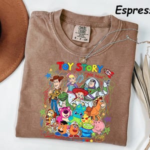 May include: Espresso-colored t-shirt featuring a colorful graphic of Toy Story characters. The design includes Woody, Buzz Lightyear, and other characters from the animated film. The shirt has a crew neck and short sleeves. A silver necklace is draped across the shirt.
