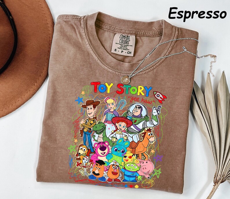 May include: Espresso-colored t-shirt featuring a colorful graphic of Toy Story characters. The design includes Woody, Buzz Lightyear, and other characters from the animated film. The shirt has a crew neck and short sleeves. A silver necklace is draped across the shirt.