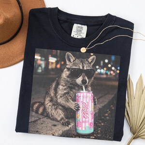 May include: Navy blue Comfort Colors t-shirt featuring a raccoon wearing sunglasses and drinking from a can of "Flav" energy drink. A gold necklace and a brown hat are also visible.