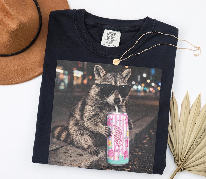 May include: Navy blue Comfort Colors t-shirt featuring a raccoon wearing sunglasses and drinking from a can of "Flav" energy drink. A gold necklace and a brown hat are also visible.