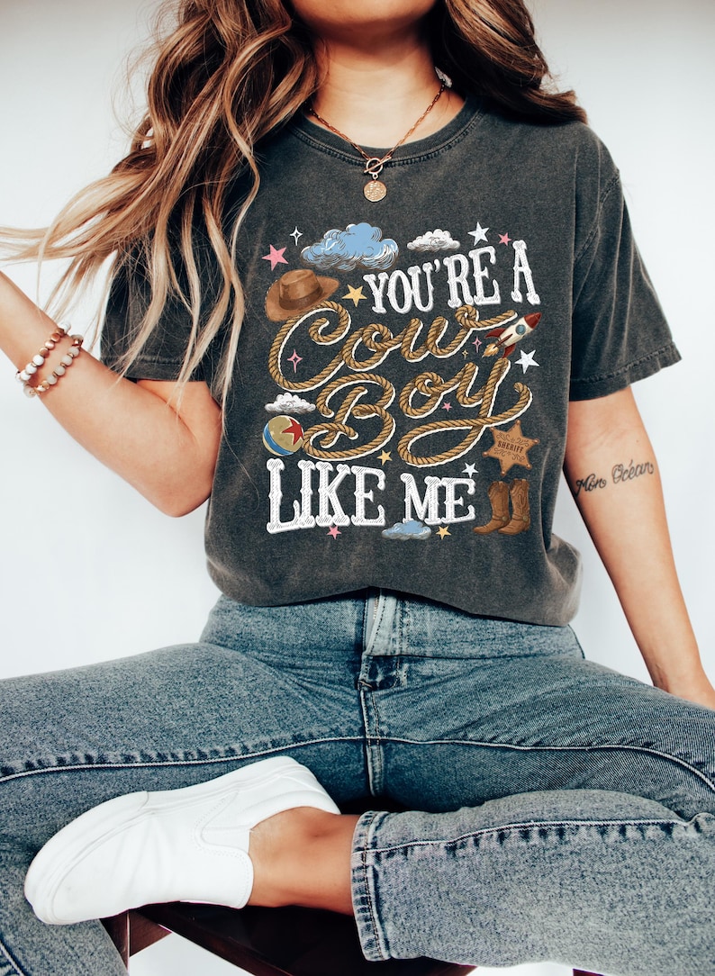 K&ouml;nnte beinhalten: Ein anthrazitfarbenes Kurzarm-T-Shirt mit einem Grafikdruck, der den Text "YOU'RE A COWBOY LIKE ME" in wei&szlig;er, seilartiger Schrift zeigt. Das Design umfasst einen braunen Hut, eine Rakete, einen Sheriffstern, Stiefel und kleine Sterne.