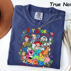 May include: True navy blue t-shirt featuring a colorful graphic of Toy Story characters. The design includes Woody, Buzz Lightyear, and other beloved characters from the animated film. The shirt has a crew neck and short sleeves. A silver necklace is draped over the shirt.
