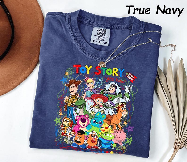 May include: True navy blue t-shirt featuring a colorful graphic of Toy Story characters. The design includes Woody, Buzz Lightyear, and other beloved characters from the animated film. The shirt has a crew neck and short sleeves. A silver necklace is draped over the shirt.