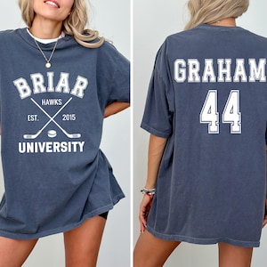 May include: A dark blue t-shirt with white text and graphics. The front features "Briar Hawks University" with crossed hockey sticks and the year "2015." The back displays "Graham" and the number "44."