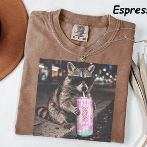 May include: Espresso-colored t-shirt featuring a graphic of a raccoon wearing sunglasses and holding a can of soda. The shirt has a crew neck and a small tag on the collar. A silver necklace is draped across the shirt.
